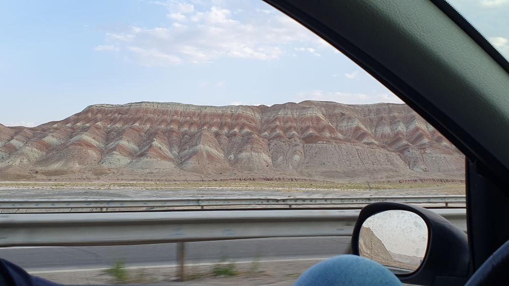 photo à travers la fenetre de la voiture sur une route en Iran montrant des dunes irisées ocres et vertes aux abords de Zanjan