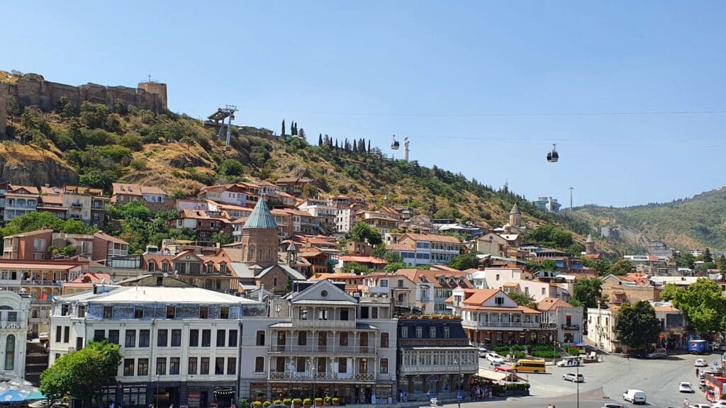 panorama du centre historique de Tbilissi avec des batisses traditionnelles au premier plan, le téléphérique et la forteresse à l'arrière plan, sous un soleil d'été